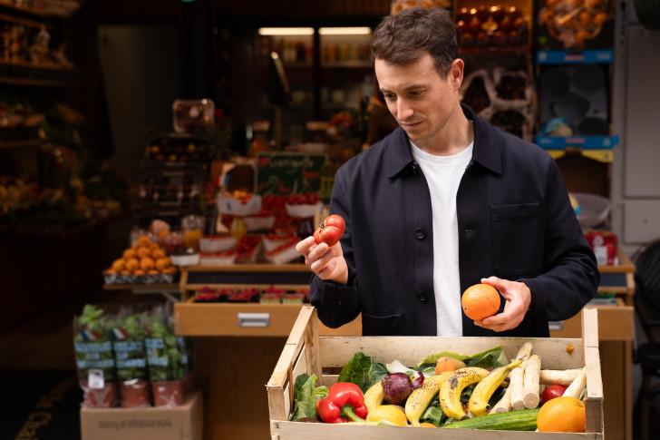 Hugo Clément regarde la tomate qu'il tient dans sa main droite. Devant lui une cagette remplie de fruits et légumes et derrière lui un étal 